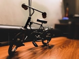 A miniature metal bicycle is placed on a wooden surface, with soft lighting casting shadows. Details of the bike, including the handlebars and wheels, are visible. The background is blurred, suggesting a focus on the bike.