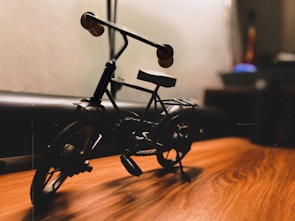 A miniature metal bicycle is placed on a wooden surface, with soft lighting casting shadows. Details of the bike, including the handlebars and wheels, are visible. The background is blurred, suggesting a focus on the bike.