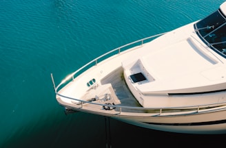 Aerial view of the bow of a white luxury yacht on calm, teal-colored water. The deck is equipped with metal railings and features minimal detailing with a sleek, modern design.