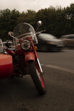 A vintage red motorcycle with a sidecar is parked on the side of a road. The motorcycle has a clear windshield and round headlight, and there are trees in the background. Cars are blurred in motion on the road, indicating movement and speed.