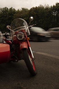 A vintage red motorcycle with a sidecar is parked on the side of a road. The motorcycle has a clear windshield and round headlight, and there are trees in the background. Cars are blurred in motion on the road, indicating movement and speed.