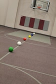 A gymnasium featuring a basketball hoop on the wall and six colorful balls aligned on the floor, including green, white, red, yellow, blue, and another white ball. The walls are padded and there is a maroon and grey padding underneath the hoop.