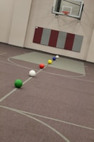 A gymnasium featuring a basketball hoop on the wall and six colorful balls aligned on the floor, including green, white, red, yellow, blue, and another white ball. The walls are padded and there is a maroon and grey padding underneath the hoop.