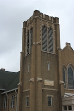 A tall, brick church tower with arched windows and louvered vents. The structure features Gothic architectural elements with pointed arches and decorative stonework. A green, gabled roof is visible, and there is a sign reading 'Turner Chapel AME Church' on the lower part of the tower.