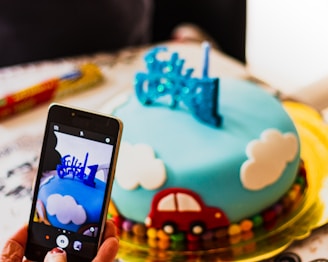 A beautifully decorated cake with a blue fondant top featuring clouds and a small red car. The cake has a number '1' and decorative elements on top. Someone is taking a picture of the cake with a smartphone.