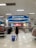 A person is walking through a retail store, pulling a blue shopping basket towards the checkout area under a sign that reads 'please pay here'. The store is bright and spacious, with shelves stocked with various items such as clothing and groceries. Numerous fluorescent lights illuminate the ceiling.