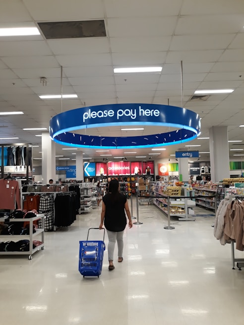 A person is walking through a retail store, pulling a blue shopping basket towards the checkout area under a sign that reads 'please pay here'. The store is bright and spacious, with shelves stocked with various items such as clothing and groceries. Numerous fluorescent lights illuminate the ceiling.