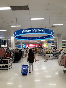 A person is walking through a retail store, pulling a blue shopping basket towards the checkout area under a sign that reads 'please pay here'. The store is bright and spacious, with shelves stocked with various items such as clothing and groceries. Numerous fluorescent lights illuminate the ceiling.