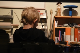 A young child with short blonde hair is seated facing away in a workspace environment. Shelves to the left and right are filled with various items, including a rolling pin, books, a figurine, and some utensils. The area looks cluttered with art supplies or tools, hinting at a creative or workshop setting.