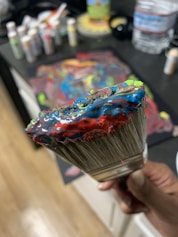 A close-up of a paintbrush covered in colorful, thick acrylic paint, including blue, red, green, and yellow. The background shows a blurred working area with various paint bottles and a painted canvas on a table surface.