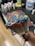 A close-up of a paintbrush covered in colorful, thick acrylic paint, including blue, red, green, and yellow. The background shows a blurred working area with various paint bottles and a painted canvas on a table surface.