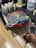 A close-up of a paintbrush covered in colorful, thick acrylic paint, including blue, red, green, and yellow. The background shows a blurred working area with various paint bottles and a painted canvas on a table surface.
