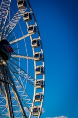 A large Ferris wheel with several enclosed cabins set against a clear blue sky. The structure's metal framework is prominently visible, and the cabins are evenly spaced along the wheel. A logo with 'RCS' is visible near the center of the wheel.