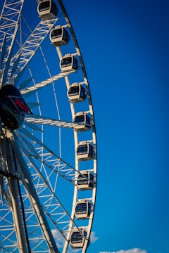 A large Ferris wheel with several enclosed cabins set against a clear blue sky. The structure's metal framework is prominently visible, and the cabins are evenly spaced along the wheel. A logo with 'RCS' is visible near the center of the wheel.