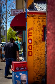 A vibrant street scene features a brightly colored yellow wall with red letters spelling 'Good Food'. A blue and red trash can is positioned on the sidewalk, adorned with stickers and graffiti. People walk along the street, and a sign above reads 'Carry-Out Here'. The setting is lively and urban with trees and other buildings visible in the background.