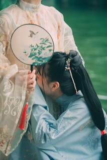 Two individuals are dressed in traditional garments, featuring intricate embroidery and delicate fabric. One person, wearing a pale robe, holds a decorative round fan with floral designs and a butterfly. The other, in a blue robe, appears to be playfully kneeling or bowing, showcasing a hair accessory.
