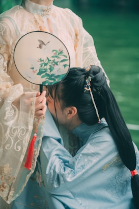 Two individuals are dressed in traditional garments, featuring intricate embroidery and delicate fabric. One person, wearing a pale robe, holds a decorative round fan with floral designs and a butterfly. The other, in a blue robe, appears to be playfully kneeling or bowing, showcasing a hair accessory.