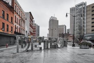 Large metallic letters spelling 'DETROIT' stand prominently in the foreground, set in an urban street with tall city buildings in the background. The scene is overcast, and the street is empty, conveying a quiet urban atmosphere.