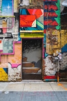 A vibrant and eclectic mural covers a brick wall, featuring a door with painted stairs that blend into the surrounding artwork. The scene consists of various abstract shapes, vivid colors, and a mix of urban and artistic elements. Stickers and urban art details are visible near the door, enhancing the street art vibe.