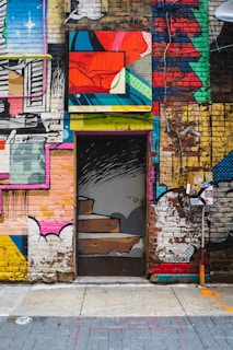 A vibrant and eclectic mural covers a brick wall, featuring a door with painted stairs that blend into the surrounding artwork. The scene consists of various abstract shapes, vivid colors, and a mix of urban and artistic elements. Stickers and urban art details are visible near the door, enhancing the street art vibe.
