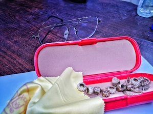 A red open glasses case lies on a dark wooden desk. In front of it are various small metallic charms or beads, possibly from a bracelet, sitting on a yellow cleaning cloth. A pair of eyeglasses is placed behind the case.