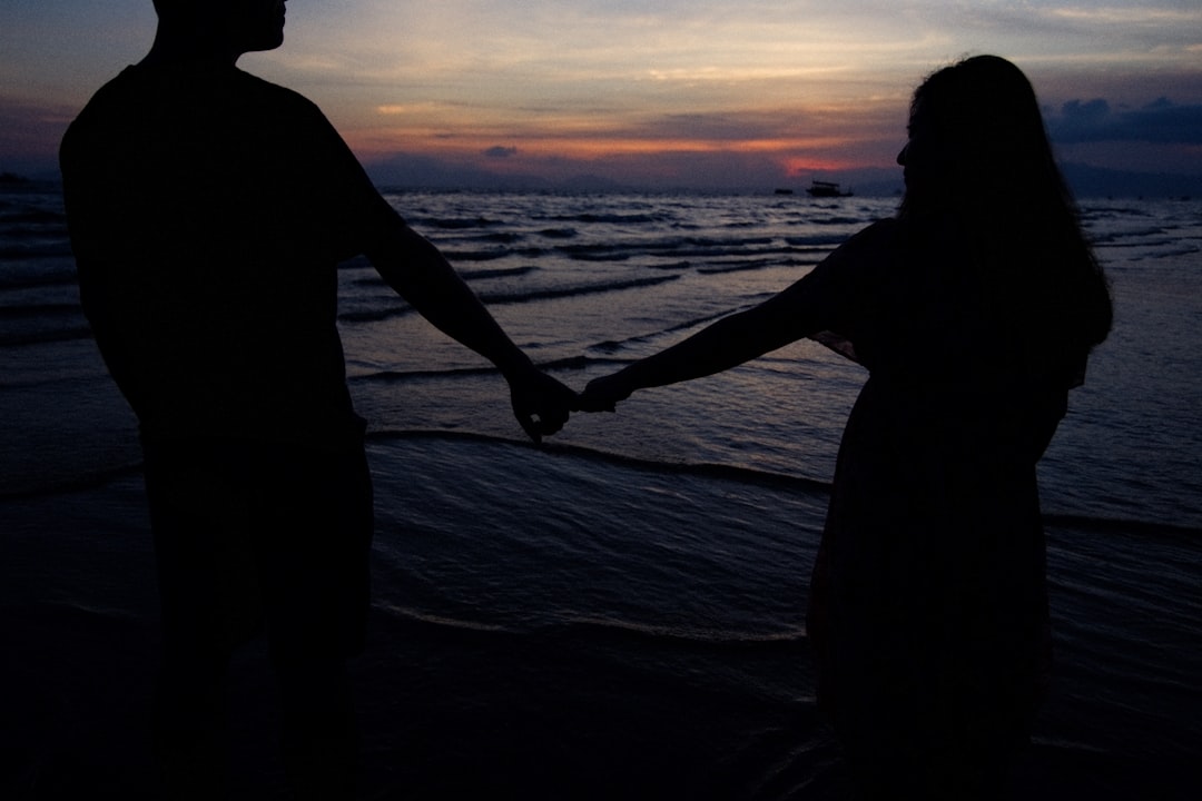 silhouette of man and woman holding hands while walking on beach during sunset