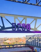 A large purple number 5280 is suspended from a metal framework against a clear blue sky. Below, an urban landscape with buildings and parking lots stretches into the distance. A mural featuring a sun is visible on a rooftop.