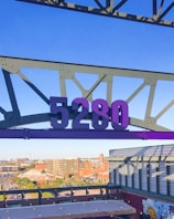 A large purple number 5280 is suspended from a metal framework against a clear blue sky. Below, an urban landscape with buildings and parking lots stretches into the distance. A mural featuring a sun is visible on a rooftop.