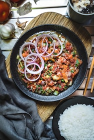 A black bowl filled with a colorful dish featuring sliced red onions, diced tomatoes, and fresh herbs, all presented on a wooden board. Accompanying the dish is a bowl of white rice, with elements of garlic and a beverage nearby. The table setting includes chopsticks and a dark cloth napkin.