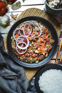 A black bowl filled with a colorful dish featuring sliced red onions, diced tomatoes, and fresh herbs, all presented on a wooden board. Accompanying the dish is a bowl of white rice, with elements of garlic and a beverage nearby. The table setting includes chopsticks and a dark cloth napkin.