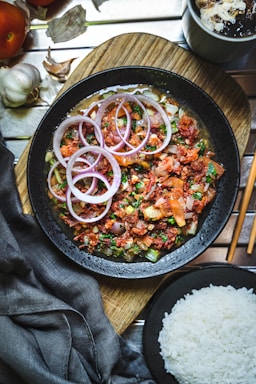 A bowl of cooked rice with fresh vegetables and herbs on a wooden table.