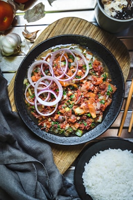 A black bowl filled with a colorful dish featuring sliced red onions, diced tomatoes, and fresh herbs, all presented on a wooden board. Accompanying the dish is a bowl of white rice, with elements of garlic and a beverage nearby. The table setting includes chopsticks and a dark cloth napkin.