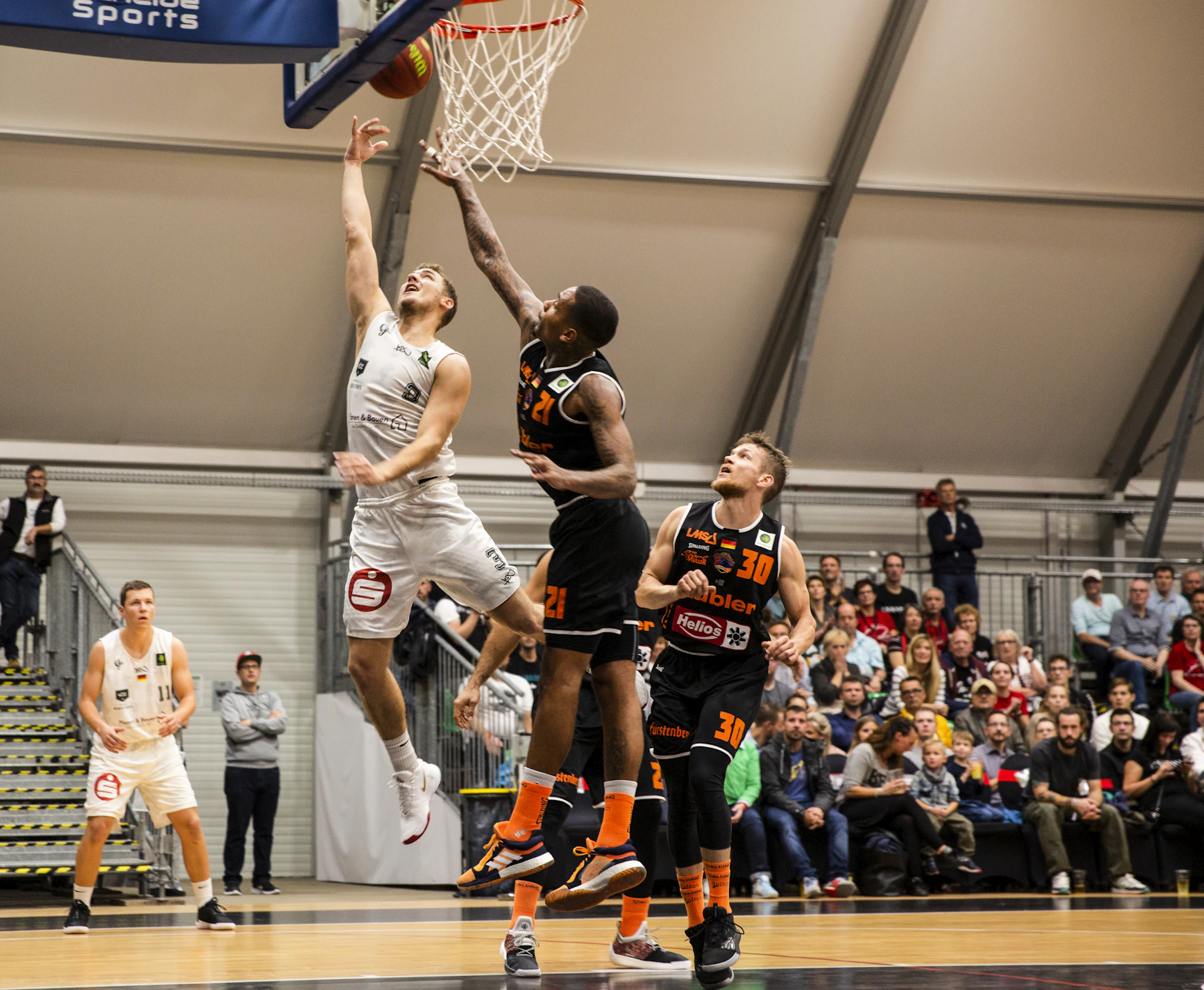 A contested layup at the rim in a small gym, spectators along the wall