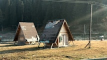 Two small, triangular wooden cabins are situated on a grassy field, with visible patches of wear and repair on their roofs. The cabins are elevated slightly above the ground and surrounded by a scenic backdrop of dense forest trees. Sunlight filters through the area, creating a serene and tranquil atmosphere.