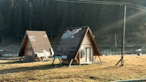Two small, triangular wooden cabins are situated on a grassy field, with visible patches of wear and repair on their roofs. The cabins are elevated slightly above the ground and surrounded by a scenic backdrop of dense forest trees. Sunlight filters through the area, creating a serene and tranquil atmosphere.