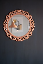 An ornate, circular mirror with an intricate wooden frame reflects a copper pendant light and blurred artwork on a light-colored wall. The mirror hangs against a dark, matte wall that contrasts with the brighter tones visible within the reflection.