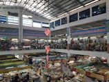 An indoor market with multiple floors is bustling with activity. Stalls are filled with various goods, including clothing, textiles, and food items. The market is crowded with people browsing and shopping. Banners with text in a foreign language are hung from the upper levels, and signs are visible throughout. The light from the high ceiling illuminates the entire space.