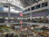 An indoor market with multiple floors is bustling with activity. Stalls are filled with various goods, including clothing, textiles, and food items. The market is crowded with people browsing and shopping. Banners with text in a foreign language are hung from the upper levels, and signs are visible throughout. The light from the high ceiling illuminates the entire space.