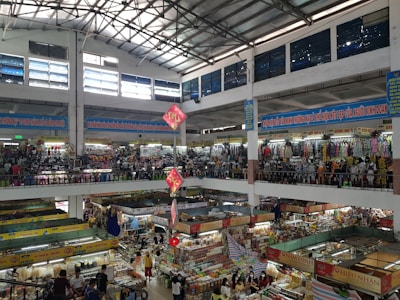 An indoor market with multiple floors is bustling with activity. Stalls are filled with various goods, including clothing, textiles, and food items. The market is crowded with people browsing and shopping. Banners with text in a foreign language are hung from the upper levels, and signs are visible throughout. The light from the high ceiling illuminates the entire space.