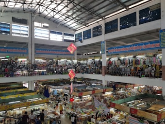 An indoor market with multiple floors is bustling with activity. Stalls are filled with various goods, including clothing, textiles, and food items. The market is crowded with people browsing and shopping. Banners with text in a foreign language are hung from the upper levels, and signs are visible throughout. The light from the high ceiling illuminates the entire space.