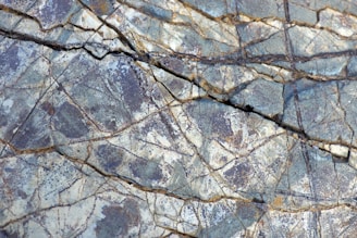An intricate pattern of natural cracks and veins in a rock surface, displaying various shades of gray and hints of brown. The texture is rough, with intersecting lines creating a network across the entire image.