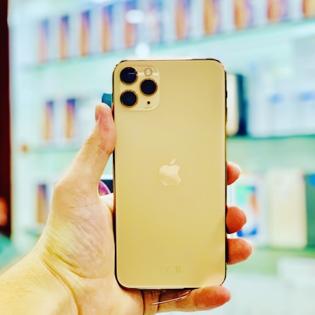 A hand holding a gold smartphone with a triple camera on the top left corner. The background appears to be a store display with blurred images of phone boxes or other products.