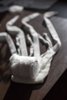 A rolled-up white fabric bandage is placed on a wooden surface, with several loose ends stretched out in different directions. The light casts soft shadows and highlights the texture of the fabric.