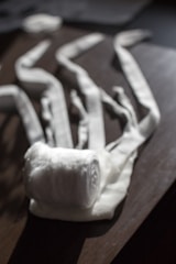 A rolled-up white fabric bandage is placed on a wooden surface, with several loose ends stretched out in different directions. The light casts soft shadows and highlights the texture of the fabric.