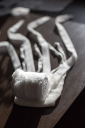 A rolled-up white fabric bandage is placed on a wooden surface, with several loose ends stretched out in different directions. The light casts soft shadows and highlights the texture of the fabric.