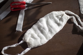 A handmade face mask made of white gauze fabric is placed on a wooden surface. Nearby, a pair of red-handled scissors and strips of fabric are visible, suggesting the mask was recently crafted.