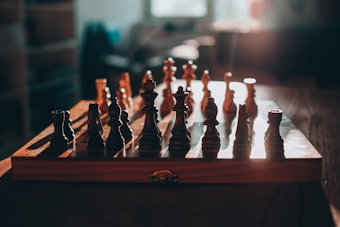 A wooden chessboard with chess pieces arranged on it, illuminated by warm, natural light. The background is softly blurred, emphasizing the chessboard as the central focus.