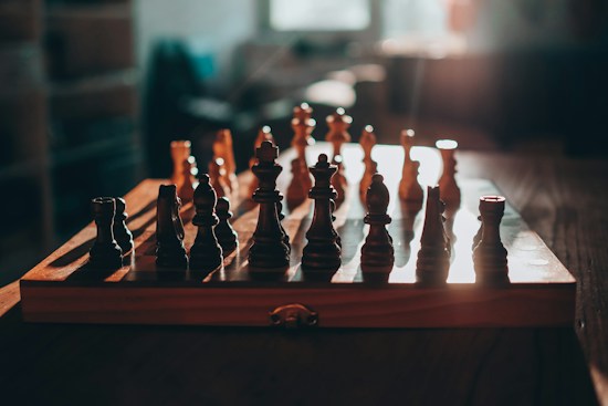 A wooden chessboard with chess pieces arranged on it, illuminated by warm, natural light. The background is softly blurred, emphasizing the chessboard as the central focus.