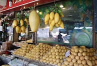 A fruit stall displays a large quantity of ripe yellow mangoes. Some mangoes are hanging in clusters above, while others are neatly stacked on a stall in front. Price tags are visible alongside some packaged goods to the left. The stall is part of a market with a glass window displaying the words 'Sticky Rice Mango'.