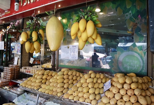 A fruit stall displays a large quantity of ripe yellow mangoes. Some mangoes are hanging in clusters above, while others are neatly stacked on a stall in front. Price tags are visible alongside some packaged goods to the left. The stall is part of a market with a glass window displaying the words 'Sticky Rice Mango'.